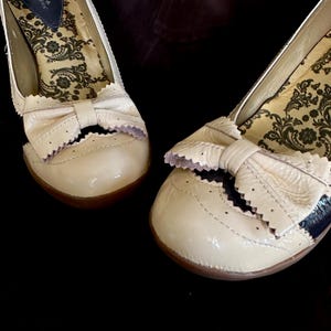 May include: Pair of cream-colored patent leather flats with bow accents. The shoes have a rounded toe and a decorative scalloped edge. The interior features a patterned fabric. The shoes are set against a dark background.