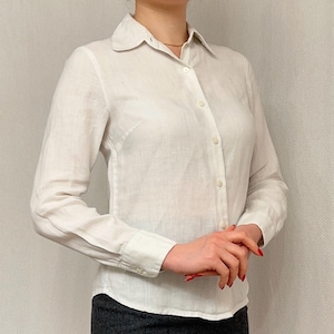 May include: A white button-down shirt with long sleeves. The shirt features a classic collar and buttoned cuffs. The fabric appears to be a lightweight linen or cotton blend. The shirt is worn by a person.
