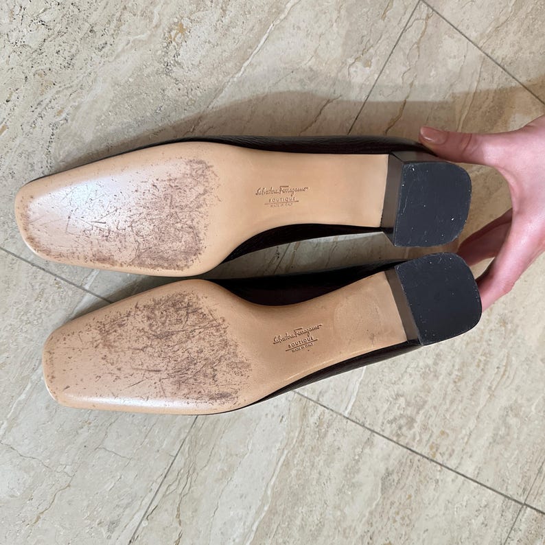 May include: A pair of dark brown leather Salvatore Ferragamo flats, with a square toe and a small, black heel. The soles are beige with wear marks, and the brand name is visible.