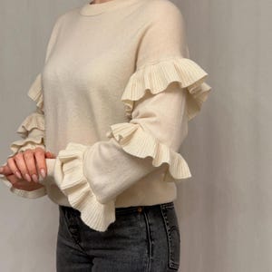 Puede incluir: Suéter color crema con mangas con volantes. El suéter tiene cuello redondo y está hecho de un material suave y texturizado. Los volantes caen en cascada por las mangas, agregando un elemento decorativo. El modelo lleva vaqueros grises oscuros.