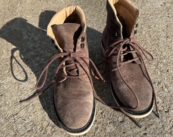 May include: A pair of brown suede boots with laces. The boots have a light-colored interior and a black and white sole. The laces are brown and tied in a bow. The boots are sitting on a concrete surface.