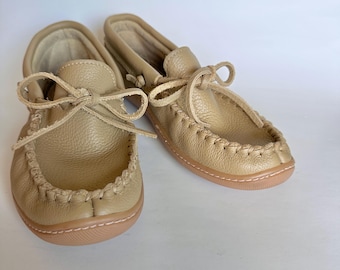 May include: A pair of tan leather moccasins with stitched detailing and laces tied in bows. The shoes have a rounded toe and a light brown sole. The moccasins are made of a soft, textured leather.