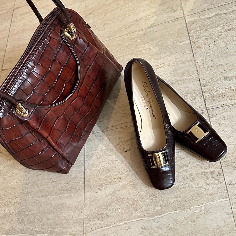May include: A brown leather handbag and matching pair of Salvatore Ferragamo flats. The handbag has a structured shape with a top handle and gold-tone hardware. The flats feature a square toe and a gold-tone buckle.
