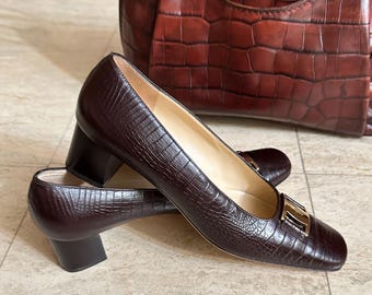 May include: Dark brown textured leather pumps with a block heel and a gold buckle. A matching brown leather tote bag is in the background. The shoes have a square toe and a low heel, approximately 5 cm.