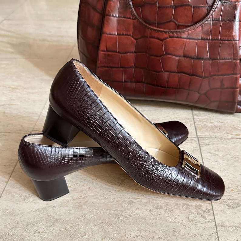 May include: A pair of dark brown, textured leather pumps with a block heel and a gold-tone buckle. A matching brown leather tote bag is in the background. The shoes have a square toe and a low heel.