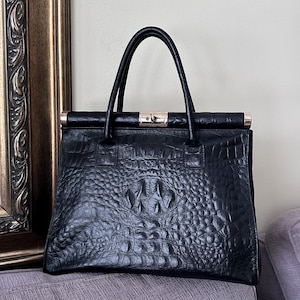 May include: Black leather handbag with a crocodile skin texture. The bag has a rectangular shape with a top handle and a gold-coloured clasp. The bag is sitting on a purple surface.