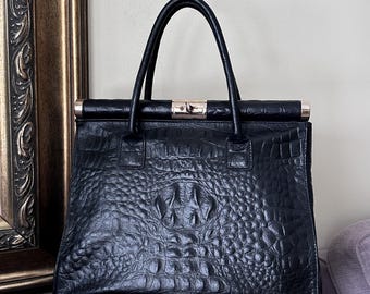 May include: Black leather handbag with a crocodile skin texture. The bag has a rectangular shape with a top handle and a gold-colored clasp. The bag is sitting on a purple surface.