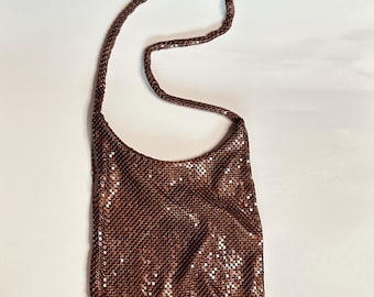 May include: A brown, metallic mesh shoulder bag with a long, braided strap. The bag has a curved top and a rectangular shape. The mesh is made of small, reflective pieces that catch the light. The bag is shown against a white background.