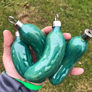 May include: Four green glass pickle baubles with silver tops. The baubles are shaped like pickles and have a shiny, glossy finish.