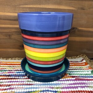 Pode incluir: Um vaso de cerâmica colorido com uma borda azul e um design listrado em tons de vermelho, laranja, amarelo, verde, azul e roxo. O vaso tem um pires correspondente.