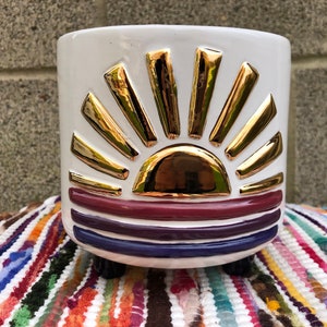 Könnte beinhalten: Weißer Keramiktopf mit einem goldenen, erhabenen Sonnenmotiv und drei violetten und burgunderfarbenen Streifen. Der Topf hat drei kleine schwarze Füße.