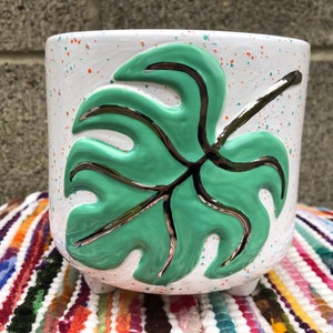 May include: White ceramic planter with a green and silver monstera leaf design. The planter has a speckled white glaze with orange and blue speckles.