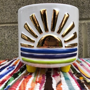 May include: White ceramic planter with a gold and blue sunset design. The planter has three yellow legs.