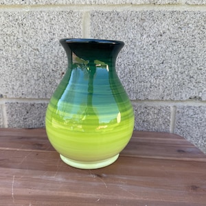 Puede incluir: Un jarrón de cerámica pintado a mano con un acabado brillante. El jarrón presenta un degradado de colores, que va desde un borde verde oscuro hasta tonos de azul y termina con una base verde lima brillante. El jarrón está sobre una superficie de madera.