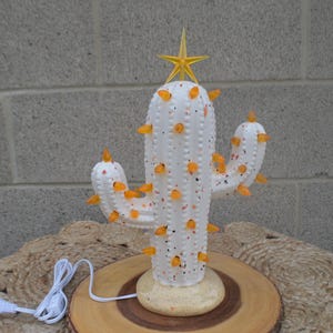 Könnte beinhalten: Weiße Keramik-Kaktuslampe mit orangefarbenen Akzenten und einem gelben Sternaufsatz. Die Lampe hat ein gesprenkeltes Design und ist mit kleinen orangen Lichtern beleuchtet. Sie steht auf einem Holzsockel mit weißem Kabel und Stecker.