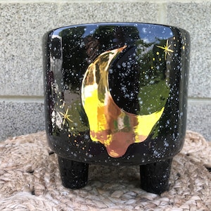 Puede incluir: Maceta de cerámica negra con un diseño de media luna dorada y un esmalte negro y blanco moteado. La maceta tiene tres patas cortas.