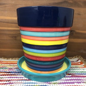 Pode incluir: Um vaso de cerâmica listrado colorido com um pires correspondente. O vaso tem uma borda azul escura e é decorado com uma variedade de cores brilhantes, incluindo vermelho, laranja, amarelo, verde, azul e roxo. O pires é de um tom azul mais claro com uma borda amarela.