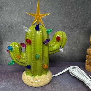 Könnte beinhalten: Ein Weihnachtsbaum aus Keramik in Form eines Kaktus mit einem gelben Sternaufsatz und bunten Lichtern. Der grüne Kaktus hat einen gesprenkelten Sockel und ein weißes Netzkabel. Eine Holzstatue von Buddha befindet sich im Hintergrund.