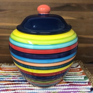 May include: A colorful ceramic jar with a lid. The jar is decorated with stripes of red, yellow, blue, green, and orange. The lid is a dark blue color with a small, round, orange knob on top.