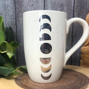 May include: White ceramic mug with a speckled finish and silver moon phases printed on the front. The mug has a large handle.