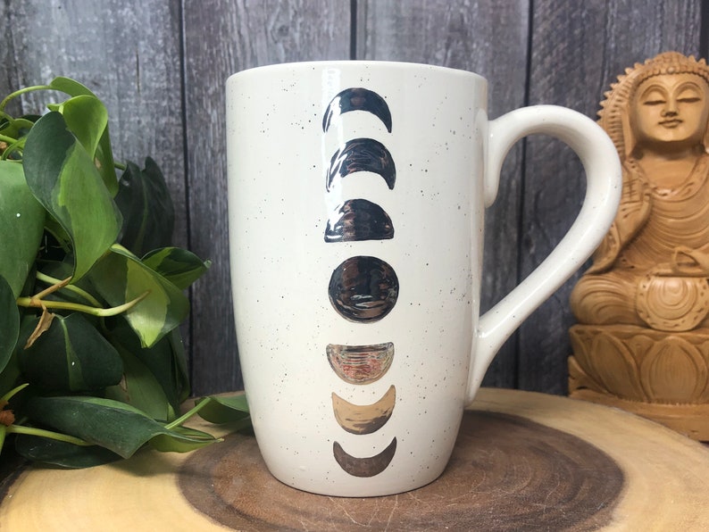 May include: White ceramic mug with a speckled finish and silver moon phases printed on the front. The mug has a large handle.