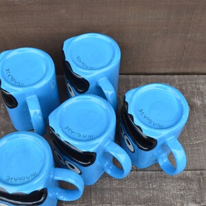 May include: Five light blue ceramic mugs with black and white cartoon face designs. Each mug has a handle and the text "INA GLAZE" on the bottom. The mugs are arranged on a wooden surface.