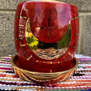 Puede incluir: Maceta de cerámica roja con un diseño de media luna y estrella dorada. La maceta tiene un platillo a juego.