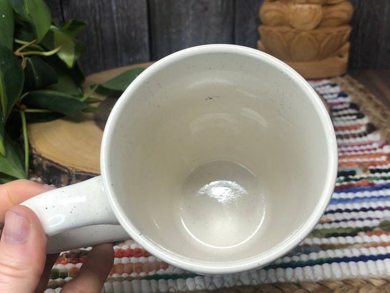 May include: White ceramic mug with a speckled finish. The mug is empty and has a handle.