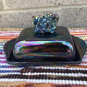 May include: A black iridescent butter dish with a crystal-like topper. The dish has a shiny, reflective surface and a rounded shape. The crystal topper is a cluster of blue and purple crystals.