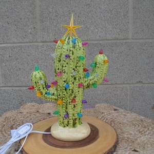 Könnte beinhalten: Ein Weihnachtsbaum aus Keramik in Form eines Kaktus mit bunten Lichtern und einem goldenen Stern als Spitze. Der hellgrüne Kaktus hat mehrere Arme und steht auf einem Holzsockel. Die Lichter sind in verschiedenen Farben, darunter Rot, Blau und Gelb.