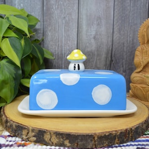 Könnte beinhalten: Eine blaue und weiße gepunktete Butterdose aus Keramik mit einem gelben Pilzdeckel. Die Dose steht auf einer Holzscheibe.