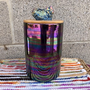 May include: An iridescent purple and green glass jar with a wooden lid and a blue crystal on top. The jar is sitting on a colourful striped rug.