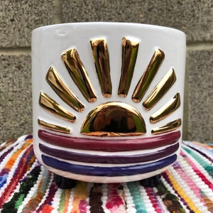 Könnte beinhalten: Weißer Keramiktopf mit einem goldenen und violetten Sonnenmotiv. Der Topf hat drei kleine schwarze Füße.