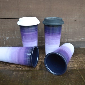 May include: Four ceramic travel mugs with a purple and white gradient design. Two mugs have white lids, and one has a dark gray lid. The mugs are arranged on a wooden surface.