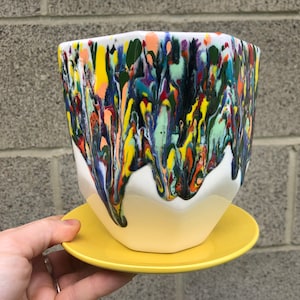 Puede incluir: Una maceta de cerámica blanca con un glaseado amarillo goteante. La maceta tiene un diseño único y abstracto con una variedad de colores, incluyendo azul, verde, amarillo, naranja y rojo.
