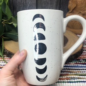 May include: White ceramic mug with a black and white moon phase design. The mug has a speckled finish and a large handle.