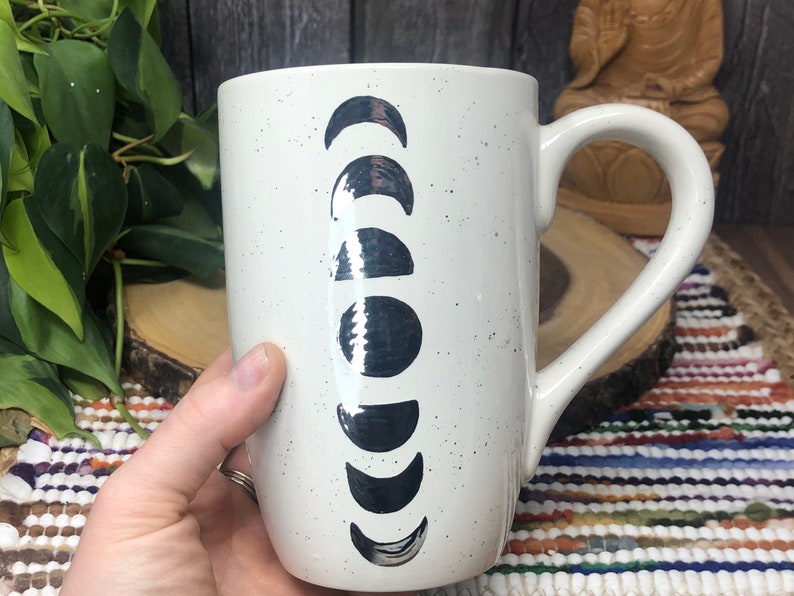 May include: White ceramic mug with a black and white moon phase design. The mug has a speckled finish and a large handle.