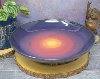 May include: A large, round, purple ceramic bowl with a swirling, ombre design in shades of pink, orange, and yellow. The bowl is sitting on a wooden slice.