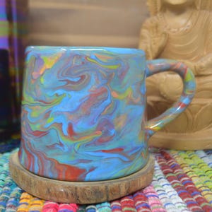 Puede incluir: Una taza de cerámica con un patrón de remolino azul, rojo, amarillo y verde. La taza se encuentra sobre un posavasos de madera. Una estatua de Buda de madera está en el fondo. La taza tiene un asa curva.