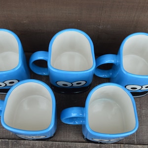 May include: Five blue ceramic mugs with a cartoon eye design. The mugs have a square shape with a white interior and a blue handle. The mugs are arranged on a wooden surface.