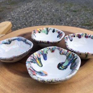 Può includere: Quattro piccole ciotole in ceramica bianca con disegni astratti di vernice a spruzzo colorata. Le ciotole sono disposte su una superficie di legno.