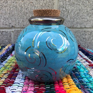 以下が含まれることがあります： 銀色の蓋とコルク栓が付いた青い陶器の壺。壺には銀色の渦巻き模様が施されています。