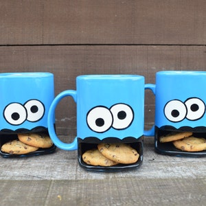 May include: Three blue ceramic mugs with a cartoon monster face design. Each mug has a black cookie pocket filled with chocolate chip cookies. The mugs have white eyes with black pupils and a black mouth.