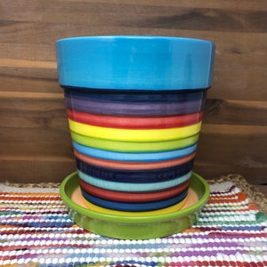 Pode incluir: Um vaso de cerâmica listrado colorido com um pires correspondente. O vaso tem o topo azul e a parte inferior verde com listras alternadas de vermelho, amarelo, roxo e azul.