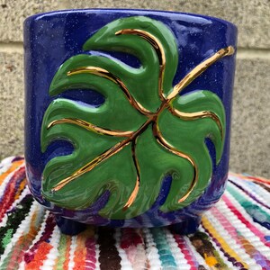 May include: A blue ceramic planter with a green and gold leaf design. The planter has three legs and is decorated with a speckled glaze.