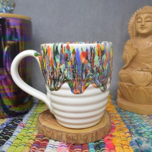 May include: White ceramic mug with a colorful, dripped glaze design. The mug sits on a wooden coaster, with a wooden Buddha statue and a decorative jar in the background. The mug has a capacity of approximately 12 ounces.