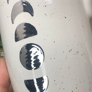 May include: White ceramic mug with a black and silver moon phase design. The mug has a speckled white glaze.