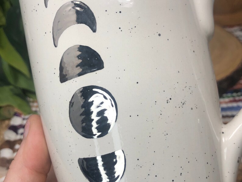 May include: White ceramic mug with a black and silver moon phase design. The mug has a speckled white glaze.