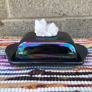 May include: A black iridescent butter dish with a white crystal on top. The dish is on a colorful striped rug.