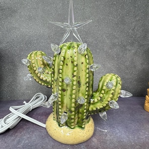 Könnte beinhalten: Ein Weihnachtsbaumaufsatz aus Keramik in Form eines Kaktus mit einem klaren Stern und Lichtern. Der Kaktus ist grün mit braunen Flecken und steht auf einem gelben Sockel.